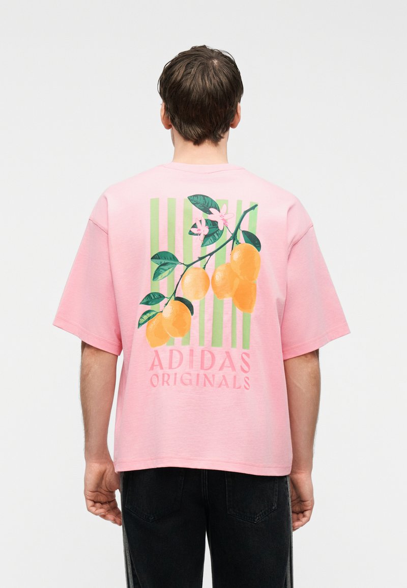 Άνδρας που φοράει ροζ μπλουζάκι Adidas Originals με κλαδί κίτρινων λεμονιών και πράσινα φύλλα πάνω σε πράσινες ρίγες στην πλάτη.