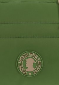 Tela de nylon verde con una textura suave presenta costuras horizontales. En el centro, un emblema redondo dice "Coronel Tapiocca Viajes y Aventura."