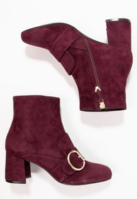 Burgundy mockaskinnankle stövlar med rund tå, klack i blockstil, sidslock, och en dekorativ gyllene spänne över framsidan.