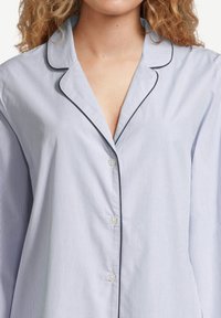 Zbliżenie na osobę w jasnoniebieskiej koszuli w paski z białymi liniami, z ciemnym wykończeniem przy kołnierzy i linii guzików.