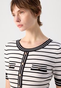 Haut en maille rayé avec un col rond, des manches courtes, des détails en boutons et deux poches poitrine avec des rayures horizontales en noir et blanc.