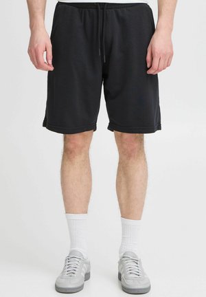 Shorts en polaire noire avec une taille élastique et un cordon de serrage, dotés d'une coupe décontractée et d'ouvertures ourlées, associés à des baskets grises et des chaussettes blanches.