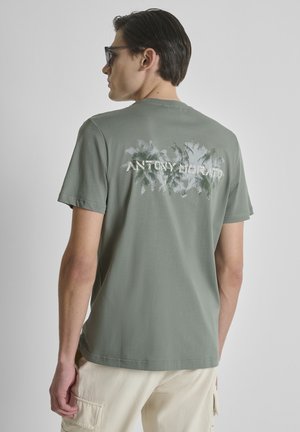 Joven con gafas de sol y camiseta verde con gráfico de palmera blanca y texto "ANTONY MORATO" en la parte trasera.