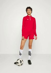 Piros sportos, hosszú ujjú felső cipzáros gallérral, piros rövidnadrággal párosítva. Fekete focicipők és csíkos fehér zoknik. Focilabda a lábánál.