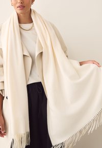 Cream-colored wool scarf with fringed edges, draped elegantly. Soft texture, lightweight design, and generous size for versatile styling.