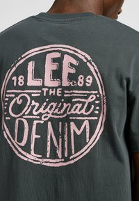 Szara bawełniana koszulka z dużym różowym okrągłym logo na plecach, na którym widnieje napis "LEE THE ORIGINAL DENIM 1889" z postarzanymi detalami.