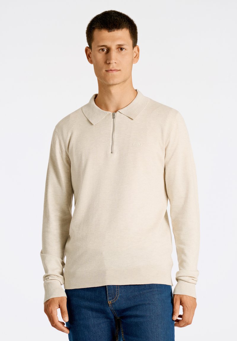 Beige pitkähihainen polo, jossa on neljänneksen vetoketju ja kaulus, jossa on teksturoitu kangas ja ribbireunat, yhdistettynä sinisiin farkkuihin.