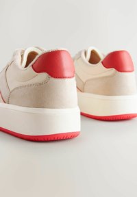 Sneaker in suede beige con un tallone in pelle rossa e suola in gomma. Il design presenta una texture liscia e dettagli di cucitura delicati.