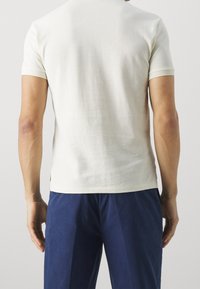 Man met een effen off-white korte mouwen shirt en marineblauwe broek, gezien vanaf de achterkant tegen een neutrale achtergrond.
