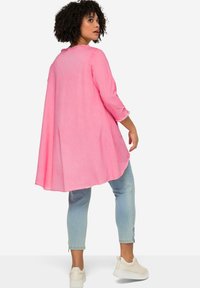 Rosa, lässige Tunika mit Dreiviertelärmeln, abgerundetem Saum und strukturiertem Stoff. Getragen mit hellblauen Jeans und weißen Sneakern.