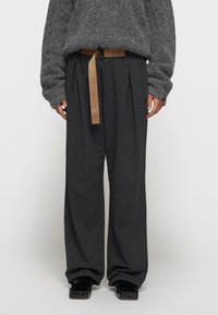 Grijze, oversized trui van textuurstof, gecombineerd met donkere, wijdvallende broek met plooien en een tan riem. Zwarte schoenen maken de look af.