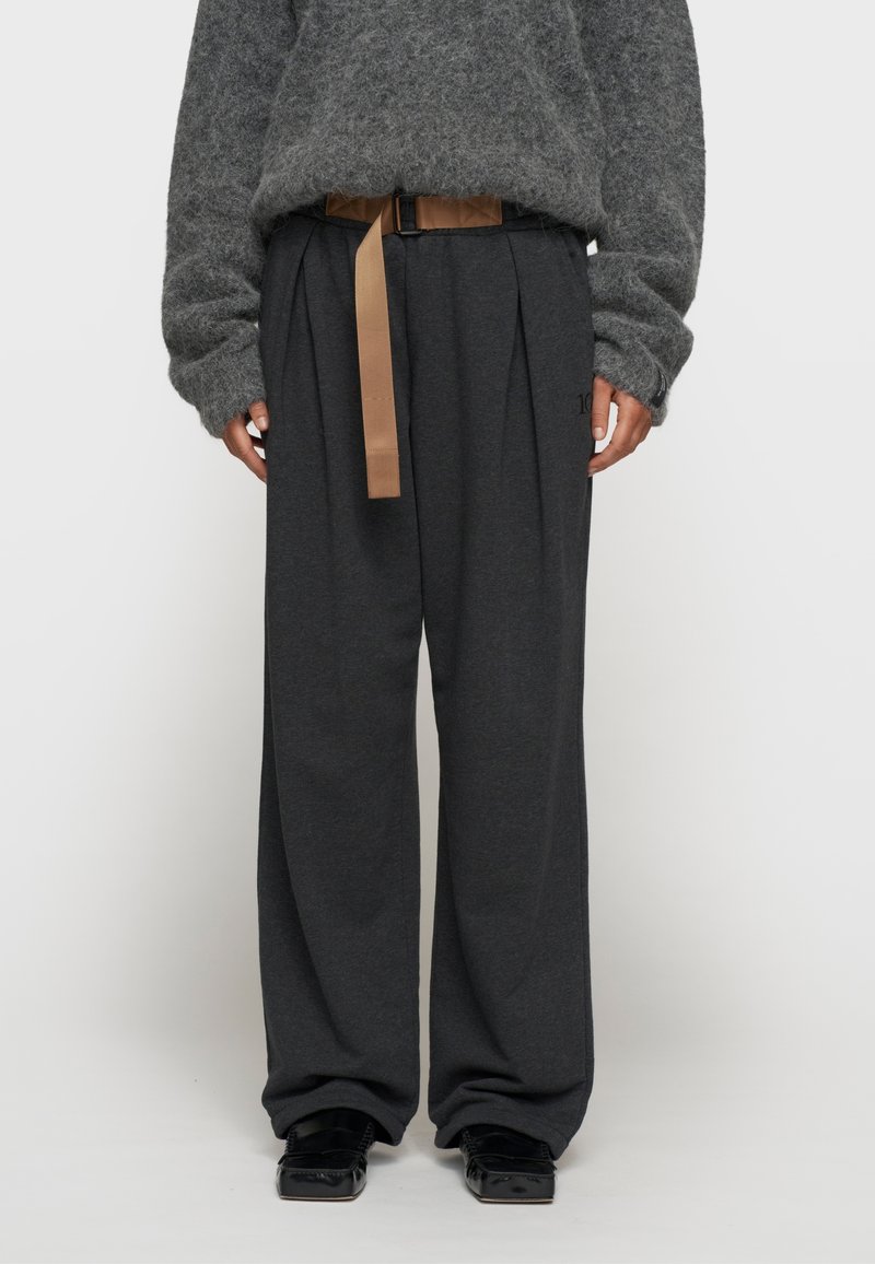 Grijze, oversized trui van textuurstof, gecombineerd met donkere, wijdvallende broek met plooien en een tan riem. Zwarte schoenen maken de look af.