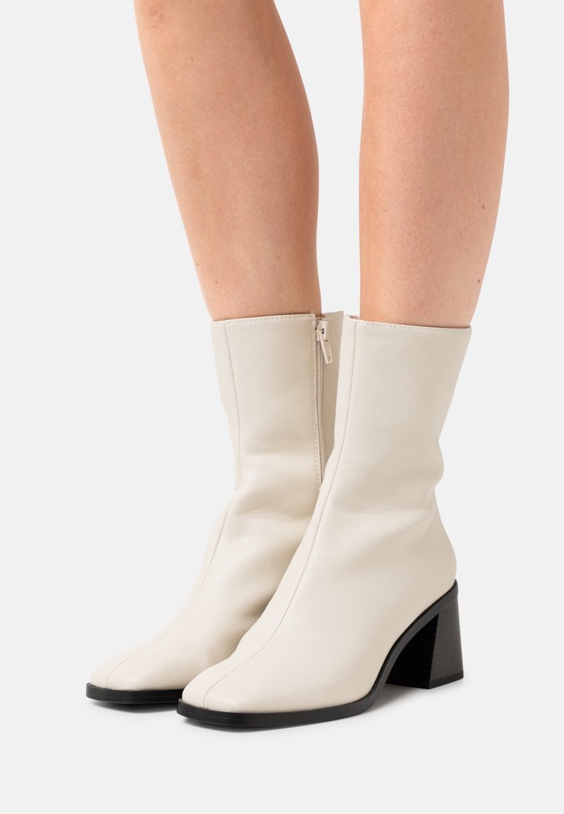 Bottes mi-mollet en cuir crème avec fermetures éclair latérales et talons carrés portées sur des jambes nues contre un fond uni.