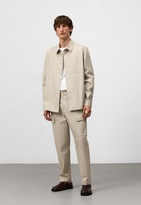 Giacca camicia in cotone beige con chiusura a bottoni, abbinata a pantaloni cargo coordinati con tasche laterali. Scarpe marroni completano l'outfit.