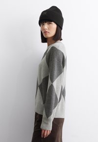 Grauer Wollpullover mit einem Rautenmuster in dunklerem Grau. Kombiniert mit einer schwarzen, gerippten Beanie für zusätzliche Wärme. Glatte Textur und lässige Passform.