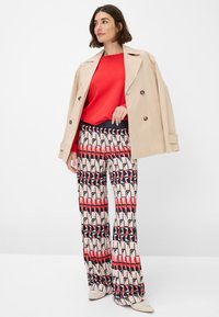 Beige trench coat over a red top, paired with patterned wide-leg pants in red, black, and white. Shoes are light-colored with rounded toes.