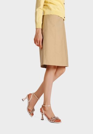 Donna che indossa una gonna beige al ginocchio, un cardigan giallo e sandali con tacco alto beige chiaro con dettagli a fiocco.