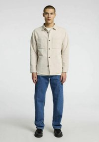 Šviesiai smėlio spalvos ilgomis rankovėmis marškiniai su dviem krūtinės kišenėmis ir juodais mygtukais, derinami su mėlynais džinsais ir juodais batais. Paprastas, struktūruotas dizainas.