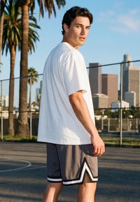 Junger Mann in weißem T-Shirt und grauer Basketball-Shorts steht auf einem Außenplatz mit Stadtskyline und Palmen im Hintergrund.