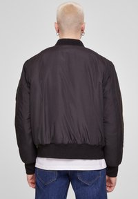 Chaqueta bomber negra con una textura suave y brillante, cuello y puños acanalados, y cintura elástica; la costura visible realza el diseño.