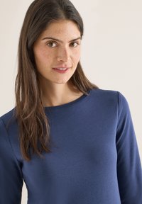 Jeune femme aux longs cheveux bruns et taches de rousseur, vêtue d'un haut à manches longues bleu marine, souriant légèrement, sur un fond clair.