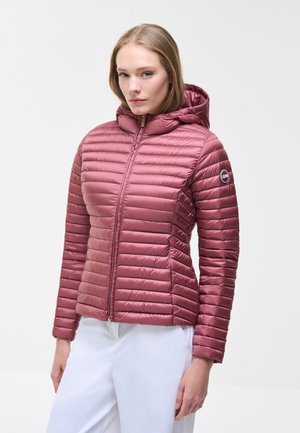 Mujer joven con chaqueta acolchada de color malva con capucha y cremallera, con parche de logo en el brazo izquierdo, combinada con pantalones blancos, de pie sobre un fondo claro.