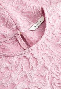 Lyserødt tekstureret stoftop med blonder-lignende design, rund halsudskæring og et lille mærke med teksten "NOELLA." Lynlåsdetalje bagpå.