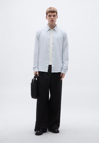 Svetlomodrá košeľa na zapínanie s jemnou textúrou, kombinovaná so širokými čiernymi nohavicami a matnými čiernymi topánkami. Drží čiernu aktovku.