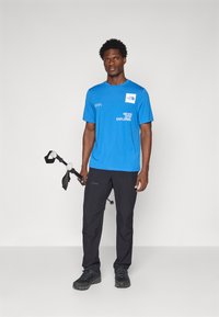 Menns blå t-skjorte med logo, slagord i hvitt, kombinert med svarte bukser og turstaver, alt mot en enkel grå bakgrunn.