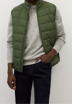 Homme portant un gilet matelassé vert par-dessus un pull en laine gris et un jean bleu foncé, avec une main dans la poche, sur un fond uni.