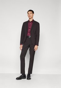 Traje oscuro con corte a medida, combinado con una camisa de botones burdeos. El modelo lleva zapatos negros y un cinturón negro. Diseño sencillo, textura suave.