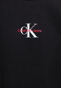 Černé bavlněné tričko s potiskem loga "Calvin Klein Jeans" v červené barvě a výrazným "CK" v bílé, umístěné ve středu hrudníku.
