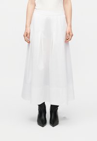 Λευκή midi φούστα ψηλοκαβαλτζέ με πιέτες, φορεμένη με μαύρα μποτάκια αστραγάλου με μυτερή μύτη, σε μονόχρωμο φόντο.