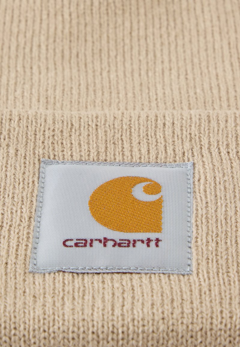 Tissu tricoté beige avec une étiquette carrée cousue montrant un logo Carhartt jaune et un texte rouge sur fond blanc.