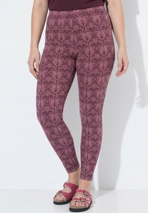 Vrouw die strakke paarse leggings met bloemenpatroon en bijpassende open sandalen met gespbandjes draagt, staand tegen een witte muur.