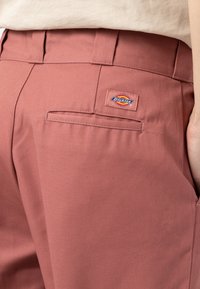 Růžové kalhoty Dickies mají hladkou tkaninu, poutka na opasek, zadní kapsu a značkovou nášivku. Design je přizpůsoben bez viditelných švových detailů.