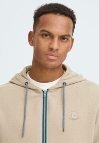 Sweat à capuche beige avec fermeture éclair, en tissu texturé, avec des accents de fermeture éclair bleus, des cordons et un logo sur la poitrine.