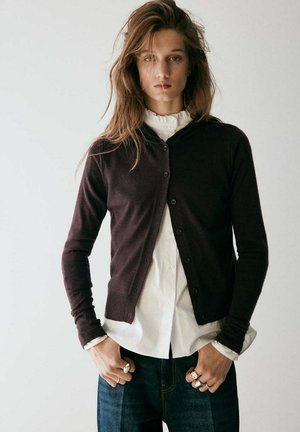 Mujer con pelo largo y castaño lleva una chaqueta de punto oscura abotonada sobre una camisa blanca con volantes y vaqueros oscuros, con anillos en los dedos.
