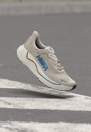 Sapatilha de corrida Hoka bege com logótipo azul, sola branca espessa, flutuando sobre asfalto com marcas rodoviárias brancas.