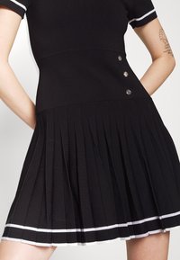 Robe noire à manches courtes avec bordure blanche aux manches et à l'ourlet, ornée de trois boutons argentés à la taille et d'une jupe plissée.