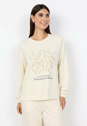 Vrouw draagt een crèmekleurige trui met een beige bladontwerp en de tekst "Paix, Amour, Bonheur Poursuis tes Rêves," glimlacht en kijkt naar de zijkant.