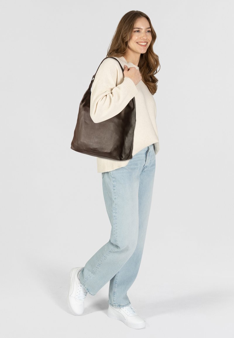 Bruine leren schoudertas met een gladde textuur, voorzien van een brede opening en subtiele branding, gecombineerd met een licht neutrale trui en blauwe jeans.