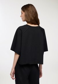 Felpa nera accorciata con maniche raglan corte, orlo grezzo e tessuto testurizzato. La vista posteriore mostra un design semplice e minimalista.