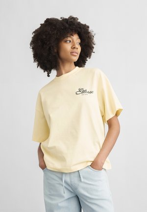 Jonge persoon met krullend haar, draagt een lichtgele oversized T-shirt met het "Ellesse"-logo en lichtblauwe broek, handen in de zakken.
