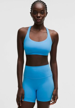Kvinde med flettet hår iført en lyseblå sports-bh og højtaljede atletikshorts mod en ensfarvet lys baggrund.