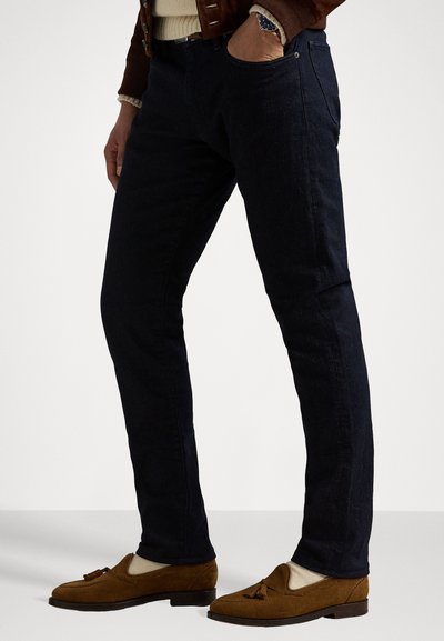 Temno modri kavbojke slim fit, z bočnimi žepi, oblečene z rjavimi semiš loafersi in svetlim puloverjem.