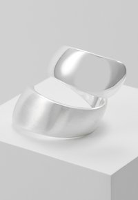 Silver metal ring with a smooth, shiny finish and a brushed texture. It features an intertwined design with a curved shape.