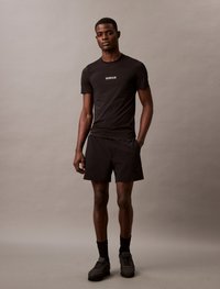 T-shirt atlético preto com mangas curtas, apresentando o logótipo branco "Calvin Klein". Combinado com calções pretos e ténis. Design simples e ajustado.