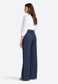 Haut blanc avec des manches bouffantes et un détail en nœud, associé à un jean bleu foncé taille haute et à jambes larges. Texture lisse, design décontracté.