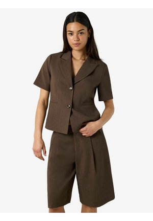 Kvinde iført brun kortærmet blazer med to knapper og matchende knælange, vide shorts, stående mod en ensfarvet baggrund.